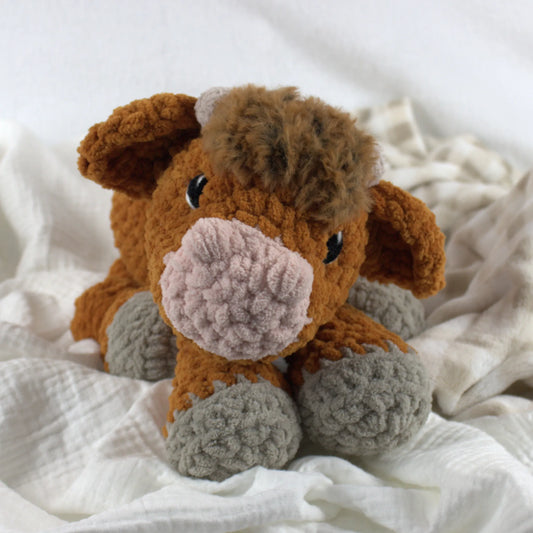 A crocheted plush cow toy with brown fur, a pink snout, and gray hooves, resting on soft white fabric.