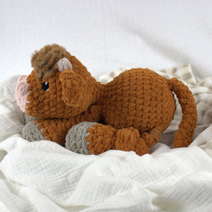 A crocheted plush toy cow in warm brown with gray hooves and a pink nose, resting on soft white fabric.