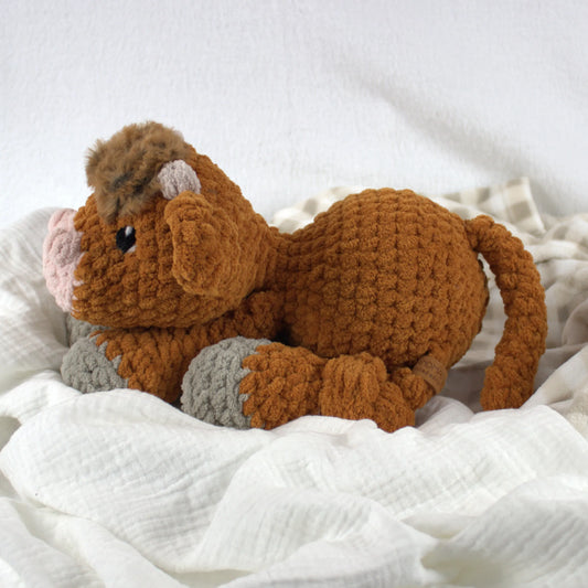 A crocheted plush toy cow in warm brown with gray hooves and a pink nose, resting on soft white fabric.