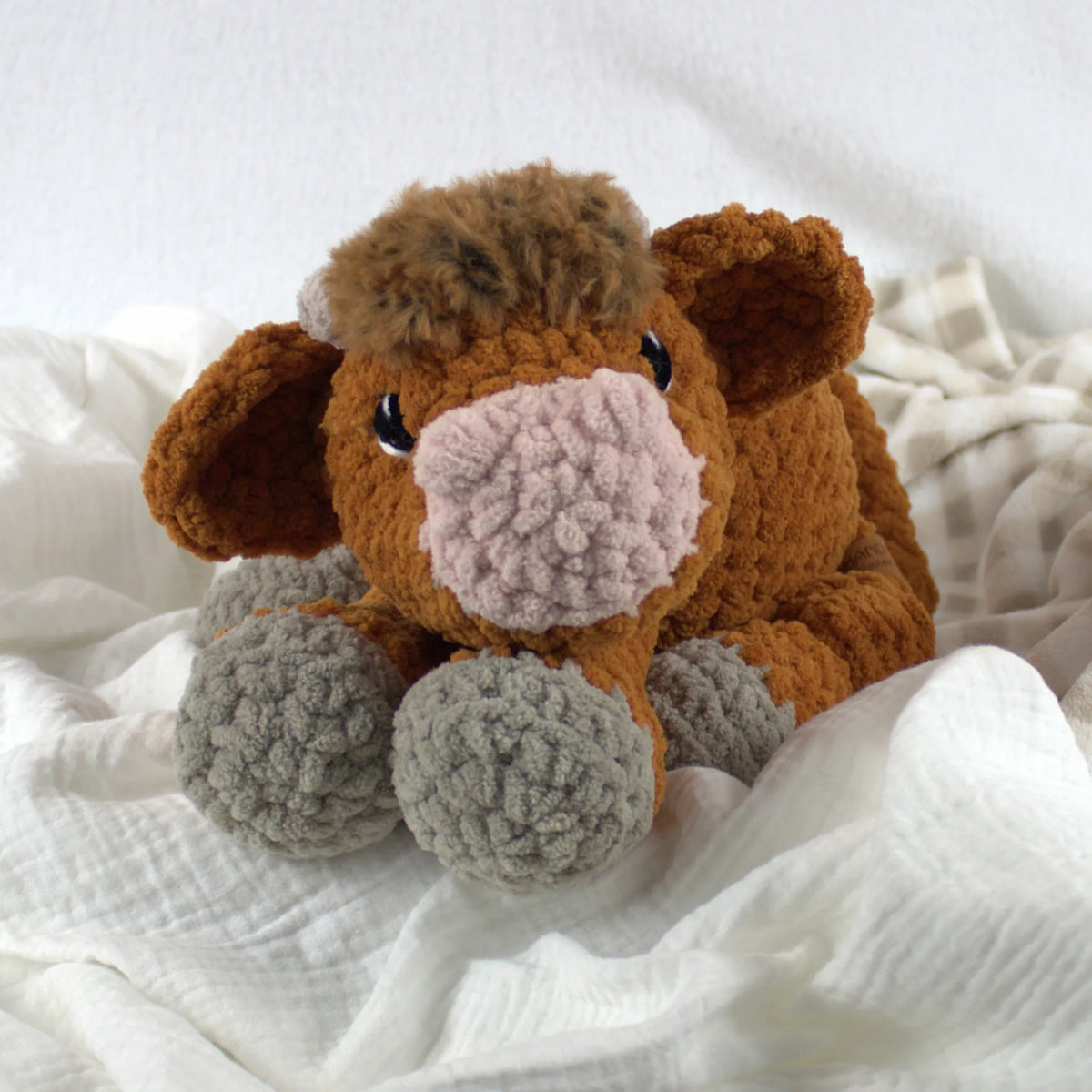 A crocheted plush cow toy with brown fur, a pink nose, and gray hooves, resting on soft white fabric.