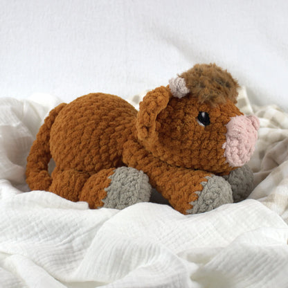 A crocheted plush toy cow in warm brown with a pink nose, black eyes, and gray hooves, resting on soft white fabric.