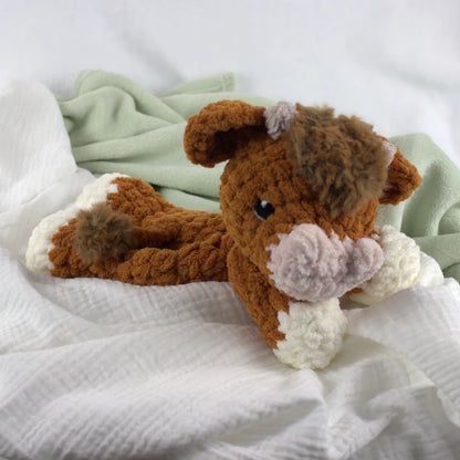 A crocheted plush cow toy in brown, white, and light pink, resting on a white textured muslin cloth with a pale green blanket in the background.