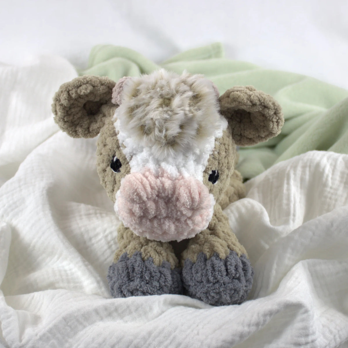 A plush crocheted cow toy with a cream and white face, pink nose, brown ears and sides, and gray hooves, resting on white textured fabric with a pale green blanket in the background.