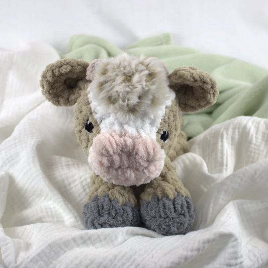 A plush crocheted cow toy with a cream and white face, pink nose, brown ears and sides, and gray hooves, resting on white textured fabric with a pale green blanket in the background.