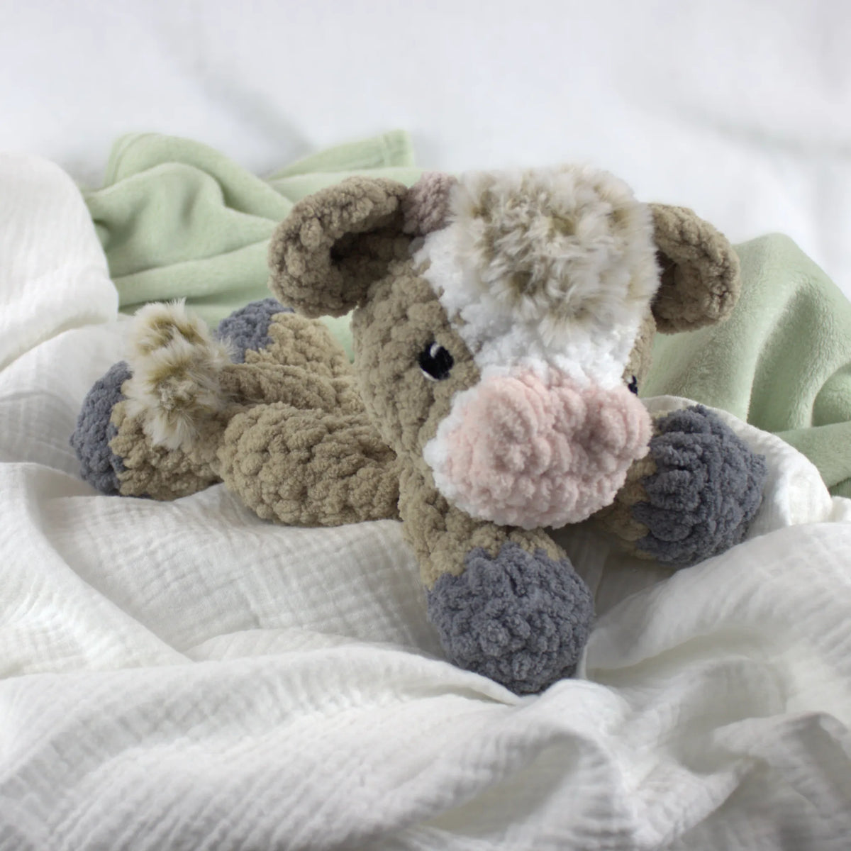 A crocheted plush cow toy lying on soft white and pale green fabric bedding. The toy has a beige body, white and cream face markings, a pink nose, and gray hooves.