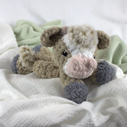 A crocheted plush cow toy lying on soft white and pale green fabric bedding. The toy has a beige body, white and cream face markings, a pink nose, and gray hooves.