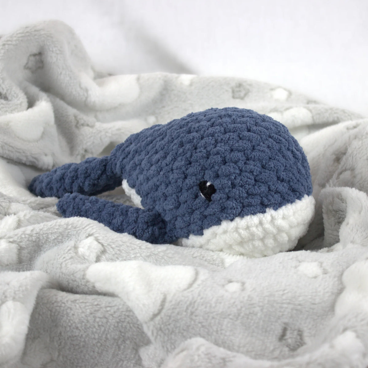 A crocheted whale plush toy in blue and white, nestled on a soft gray blanket with white cloud patterns.