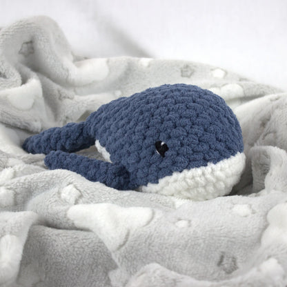A crocheted whale plush toy in blue and white, nestled on a soft gray blanket with white cloud patterns.