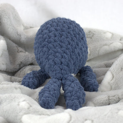 A blue crocheted whale plush toy resting on a soft white blanket with a subtle cloud pattern.