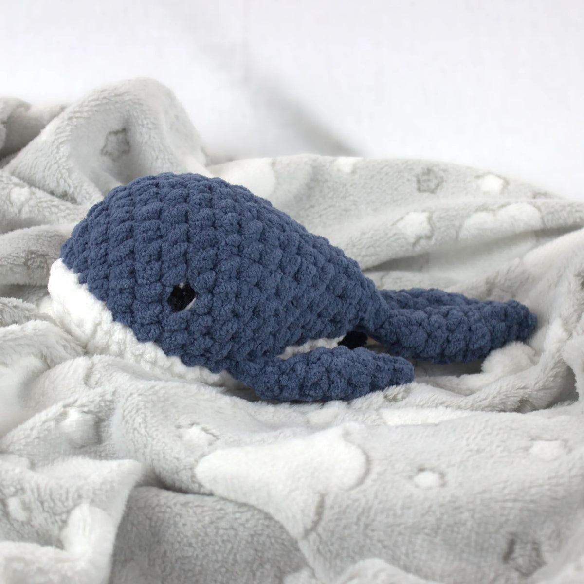 A crocheted whale plush toy in blue and white, resting on a soft gray blanket with white cloud patterns.