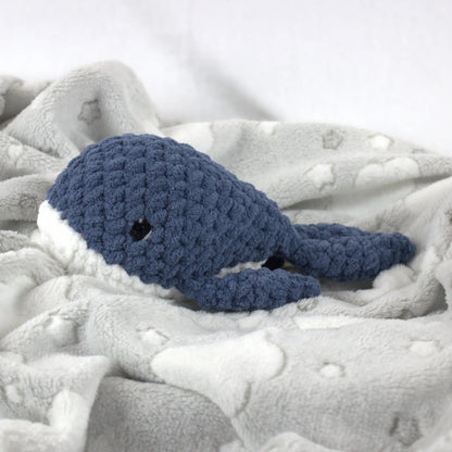 A crocheted whale plush toy in blue and white, resting on a soft gray blanket with white cloud patterns.