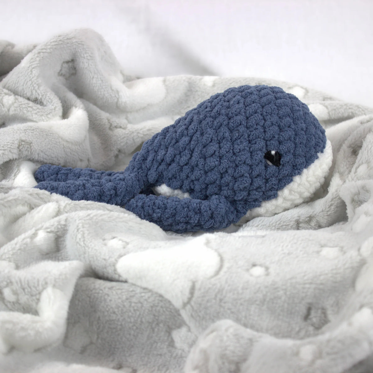A crocheted whale plush toy in blue and white, nestled on a soft white blanket with white cloud-like patterns.