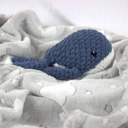 A crocheted whale plush toy in blue and white, nestled on a soft white blanket with white cloud-like patterns.